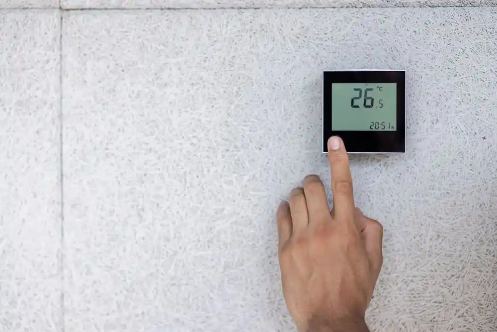 Duvara monte dijital termostat 26,5 °C gösteriyor; bir el ekrana dokunuyor